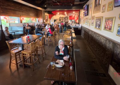 A woman sits alone at a high-top table in a busy restaurant. Other diners are seated at tables, and some are lining up at the counter. The walls feature various framed pictures. A neon sign reads Tex and Trails is visible on the floor.