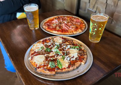 Two pizzas on a wooden table, each on a tray. One pizza has mozzarella and basil, the other has pepperoni and bacon. Two glasses of beer are beside them. A person in a black jacket sits in the background.