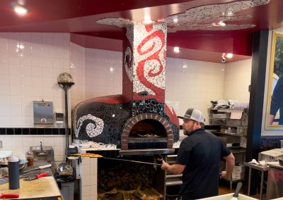 A person wearing a cap is preparing food in a kitchen with a prominent stone pizza oven adorned with mosaic art. The oven has red and white swirl designs. A portrait of a man is on the wall to the right. Various kitchen tools are visible.