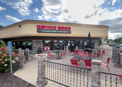 Outdoor view of Dough Bros Woodfire Kitchen, featuring a beige building with a red and black sign. Theres a patio area with red chairs and tables under umbrellas, surrounded by a black metal fence. A blue sky and clouds are visible above.