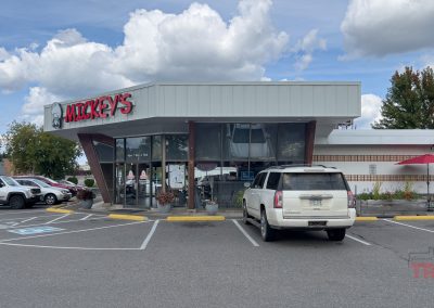 A mid-century modern style building houses Mickeys restaurant, featuring large windows and a red and white sign. Five cars are parked outside on a cloudy day. A red patio umbrella and plant beds are visible near the entrance.