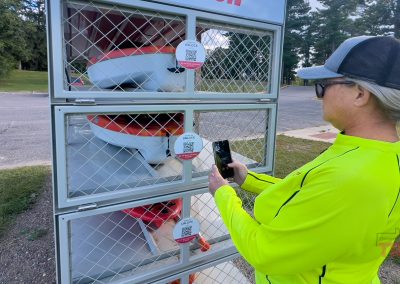 A person in a bright yellow jacket and hat uses a smartphone to scan a QR code on a life kayak rental station. The station has several compartments with life jackets inside, situated outdoors near a parking lot and trees.