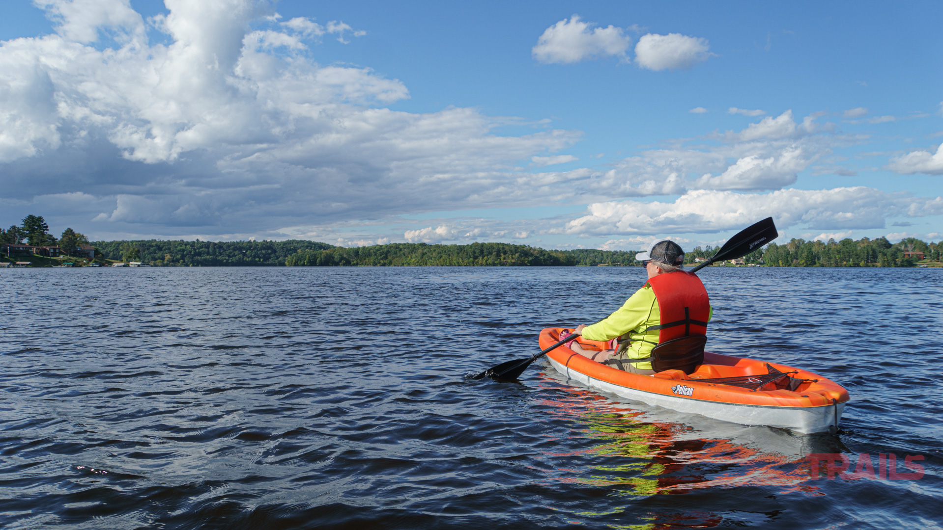 Rice Lake Brainerd A person in a bright yellow jacket and cap is kayaking on a calm lake under a partly cloudy sky. The distant shoreline is lined with trees, and the water reflects the blue of the sky and the white clouds.