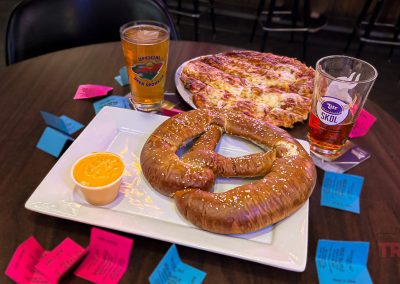 A table with a large pretzel and cheese sauce on a square white plate, a pepperoni pizza, and two glasses of beer. Colorful raffle tickets are scattered on the table.