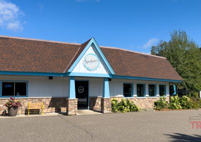 A small building with a brown shingled roof and blue trim. The sign reads Sweetwater. There are windows along the front and potted flowers by the entrance. A board with open is displayed in one window. Trees are in the background.