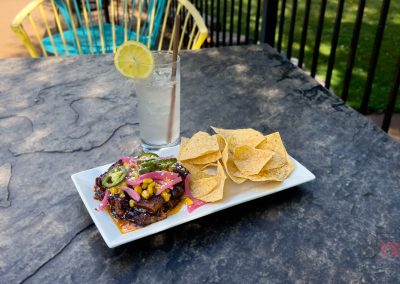 A rectangular white plate with nachos topped with jalapeños, corn, and pickled onions next to a pile of plain tortilla chips. A glass of lemonade with a lemon slice garnish is beside the plate on a stone table. A patio with a chair is in the background.