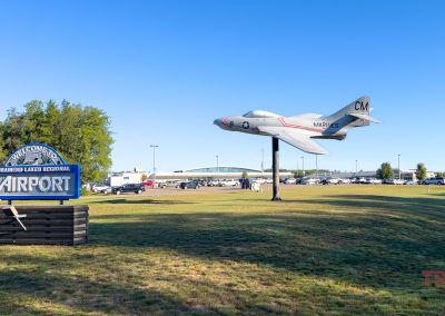 Replica of a military jet mounted on a pole at Brainerd Lakes Regional Airport. A welcome sign is in front of the aircraft. Cars and buildings are visible in the background under a clear blue sky.