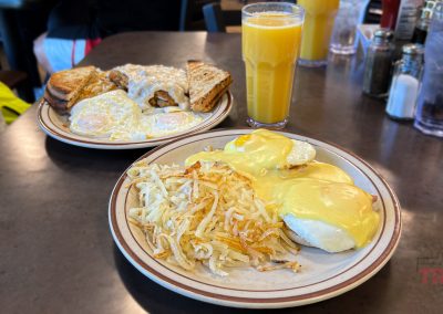Plates of breakfast food on a table: one with eggs Benedict and hash browns, the other with toast, eggs, and sausage gravy. Accompanied by a glass of orange juice and water glasses in the background.
