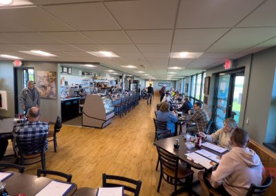 A narrow restaurant with wooden floors and ceiling lights. Diners sit at tables on the left and along a bar on the right. Large windows are on the right, providing natural light. People are engaged in conversation and eating.