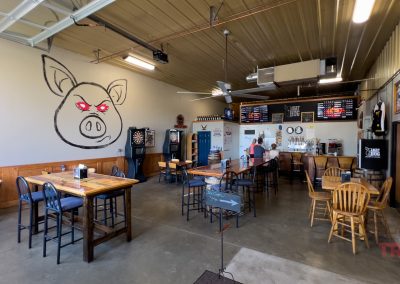 Interior of a barbecue restaurant with wooden tables and chairs, a large pig mural on the wall, and menus displayed above the counter. A person stands at the counter. The space features concrete floors and ceiling fans.