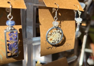 Close-up of handmade earrings displayed on brown cards. The left earring features blue flowers and the right showcases white flowers, both encased in metal frames with a decorative bead on top. Sunlight casts shadows on the display.