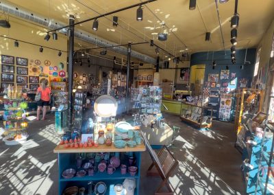 A spacious art gallery with sunlit walls displaying colorful artwork. Shelves and tables exhibit various art pieces and pottery. Two people are browsing the collection. The room has spotlights and a high ceiling with exposed pipes.