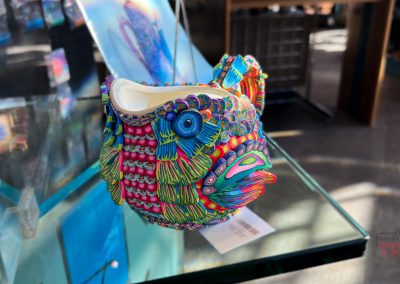 A colorful ceramic fish vase with intricate, vibrant patterns and textures is displayed on a glass shelf. The fish has prominent eyes and an open mouth, set against a backdrop of other items on display.