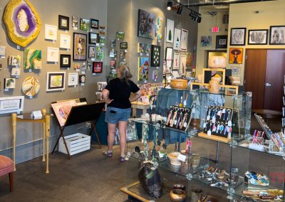 A person stands in a gallery-style shop, observing artwork displayed on the walls. Various art pieces, including paintings and crafts, are showcased. Glass display cases contain small items. The room is well-lit and decorated with vibrant colors.
