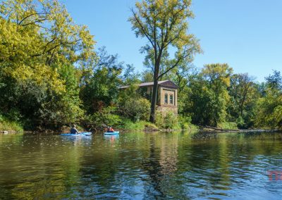 Two people kayak on a calm river surrounded by lush green trees under a clear blue sky. A stone building with arched windows is partially covered by foliage on the riverbank.