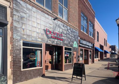 Storefronts of Dustys Bar & Lounge and adjacent shops on a sunny street, showcasing brick and tiled facades. Theres a streetlamp and visible reflections on windows. Signs and posters are visible on the windows.