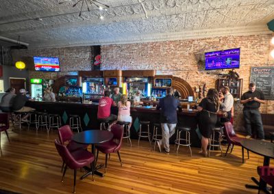 People are seated at a bar in a dimly lit room with exposed brick walls and a wooden floor. TV screens are mounted above the bar. A neon TRAILS sign is visible on the right. Tables and chairs fill the foreground.
