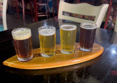Four small glasses of beer sit on a wooden serving board, showcasing a gradient from light to dark amber. The setting is a rustic bar with wooden chairs, and theres a logo reading TRAILS in the corner.