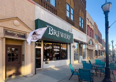 Street view of a small-town brewery with a green awning labeled Brewery, next to various storefronts. Patio tables and chairs are set up on the sidewalk under lampposts. A white flag and blue skies enhance the scene.