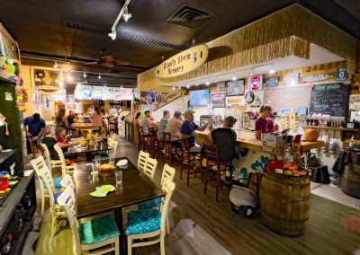A lively brewery interior with patrons seated at the bar and tables. The space is decorated with beach-themed signs, a straw-covered ceiling section, and colorful wall art. Shelves display various items, and the atmosphere is casual and inviting.