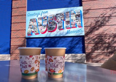 Two paper cups with autumn-themed designs sit on a table in front of a brick wall. A colorful mural reads Greetings from Austin, Minnesota with autumn leaves casting shadows on the wall.