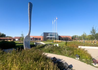 Modern building with a large glass facade and three flagpoles in front, surrounded by greenery. A tall, abstract silver sculpture is in the foreground. Clear blue sky in the background.