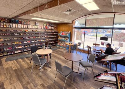 Interior of a comic book store with shelves of comics along the walls. Two people seated at separate tables are reading. Large front windows let in natural light, and chairs are arranged casually around tables in the center.