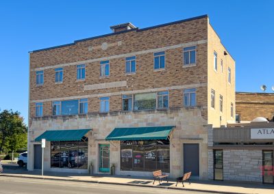 A three-story brick building with large windows. The ground floor features awnings over storefronts. Two benches are on the sidewalk. A white car is parked nearby. Blue sky and trees are visible in the background.