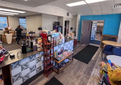 A cozy cafe interior with a counter displaying snacks and drinks. A person stands behind the counter, and another person sits on the right using a phone. The walls are painted blue and gray, and there are shelves with various items.