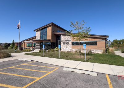 A modern building with wood and stone exterior is set under a clear blue sky. An American flag flies nearby. Accessible parking spaces line the front, and a TRAILS logo is visible in the bottom right corner.