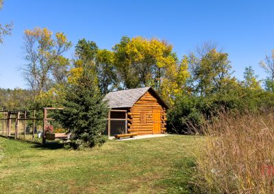 A small log cabin with a wooden fence sits in a grassy clearing surrounded by trees with green and yellow leaves under a clear blue sky. A path leads to the cabin entrance.