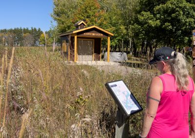A person in a pink tank top, with a ponytail and sunglasses on their head, looks at an information board on a trail. In the background, a wooden shelter sign reads Sugar Shack surrounded by tall grass and trees.