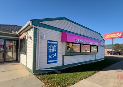 Small diner with white siding and green trim, featuring a pink awning reading Kennys Oak Grill. A blue sign on the side says Spam Town USA. A sign for an auto parts store is visible in the background.