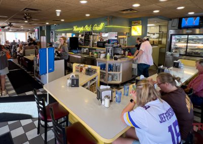 Inside a bustling diner, patrons sit at a counter and in booths. The decor is vintage with signs and menu boards. A counter displays condiments and napkins. The atmosphere feels lively and casual, with customers chatting and ordering food.