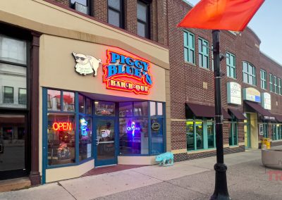 Street view of a brick building featuring Piggy Blues Bar-B-Que with neon signs, including an OPEN sign. A playful pig mascot is displayed above the entrance. Other businesses are adjacent, and a flag waves from a nearby lamppost.