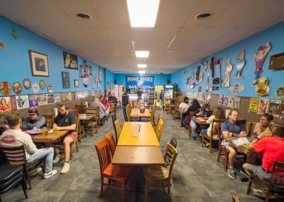 A busy restaurant with blue walls adorned with various signs and wall art. Patrons sit at wooden tables engaged in conversation. The space has a casual atmosphere, with a mixture of bright and soft lighting.