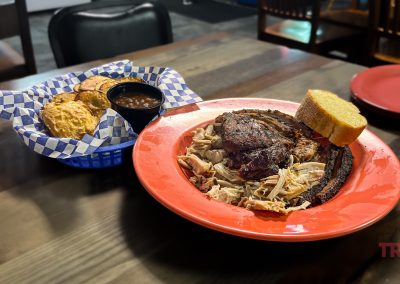 A plate of barbecue with pulled pork, ribs, and grilled meat topped with a slice of bread. A side basket contains biscuits with a small cup of sauce. The table setting includes a person in an orange shirt and wooden chairs.