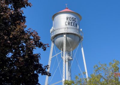 Water tower labeled Rose Creek stands against a clear blue sky, surrounded by the tops of leafy trees.