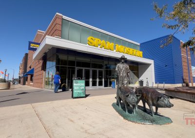 Street view of the SPAM Museum featuring a modern brick building with large glass windows. In front, a bronze sculpture depicts a person with two pigs. Clear blue sky above.
