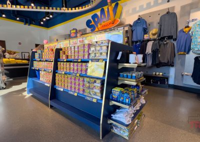 A store display featuring various SPAM products in colorful packaging. Shelves are filled with cans and boxes, surrounded by SPAM-themed merchandise such as clothing on hangers. Bright, modern interior with overhead lighting.