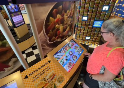 A woman in a pink shirt is using a touchscreen kiosk with food images displayed. She is standing in front of a wall filled with stacked canned goods. The setting has vibrant colors and a patterned floor.