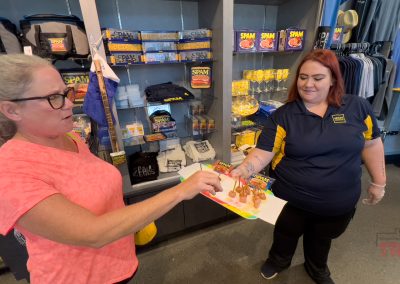 A woman wearing a pink shirt samples a snack offered by another woman in a blue and yellow uniform. They are inside a store with shelves displaying various SPAM products, apparel, and merchandise.