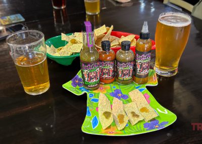 Two glasses of beer beside bowls of tortilla chips, plates of chips with hot sauce, and four bottles of hot sauce on a colorful tray. The setting is a dark, glossy table.