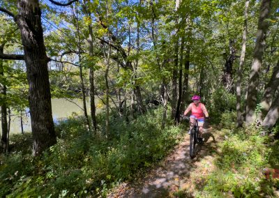 A person wearing a pink helmet and tank top rides a mountain bike on a narrow, wooded trail alongside a river. The trail is surrounded by lush green trees and foliage under a clear blue sky.