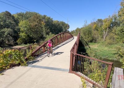 A person in a pink outfit rides a bicycle across a wooden and metal bridge surrounded by green trees and foliage on a sunny day. The path leads into a lush forested area.