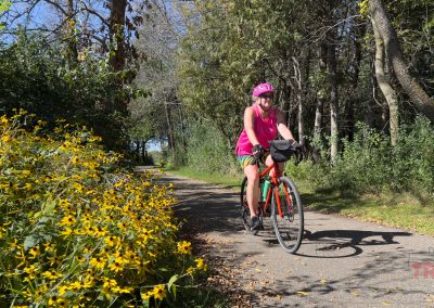 A person wearing a pink helmet and top rides a bicycle on a paved path through a wooded area. Yellow wildflowers line the path under a partly cloudy sky.