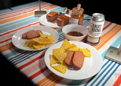 Two plates with sliced meat and tortilla chips rest on a striped tablecloth. Nearby, theres a jar of salsa, a small bowl of sauce, a can labeled Angry Hog, and a plate with two meat squares.