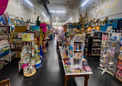 A colorful bookstore with a variety of books displayed on shelves and tables. Brightly decorated walls display artwork. Various sections showcase childrens books, novels, and graphic novels. The store is vibrant and inviting.
