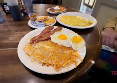 A hearty breakfast with sunny-side-up eggs, crispy bacon, and hash browns on a plate. In the background, pancakes with butter and syrup, and an omelet with a side of biscuits are visible.