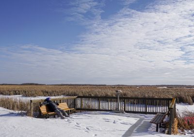 A woman wearing winter clothes lounges on a bench at an observation deck at a frozen lake